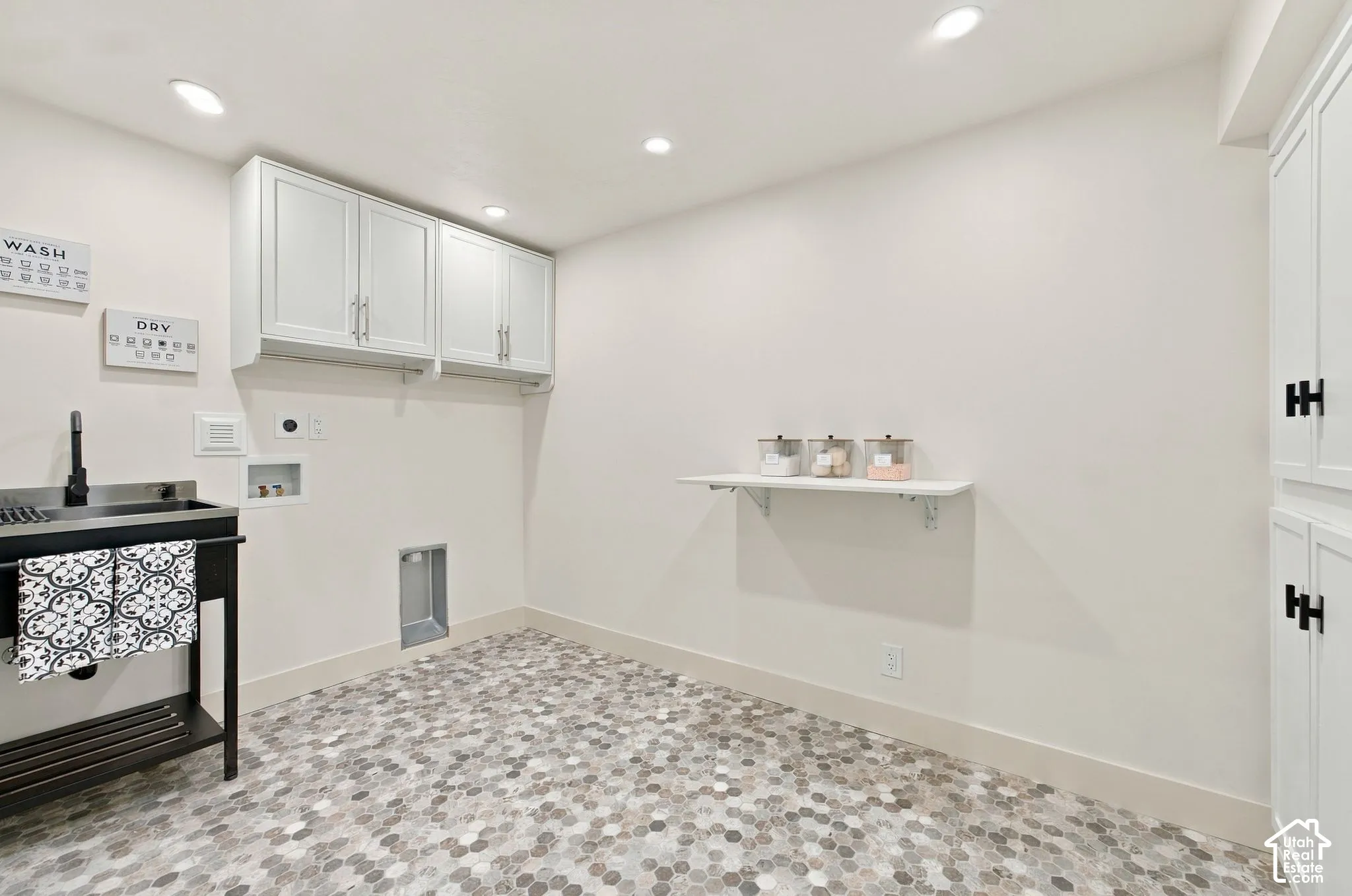 Dedicated laundry room featuring white cabinetry with ample storage, a black front-load washer and dryer unit, mosaic tile flooring, recessed ceiling lighting, and a wall-mounted shelf for supplies and organization