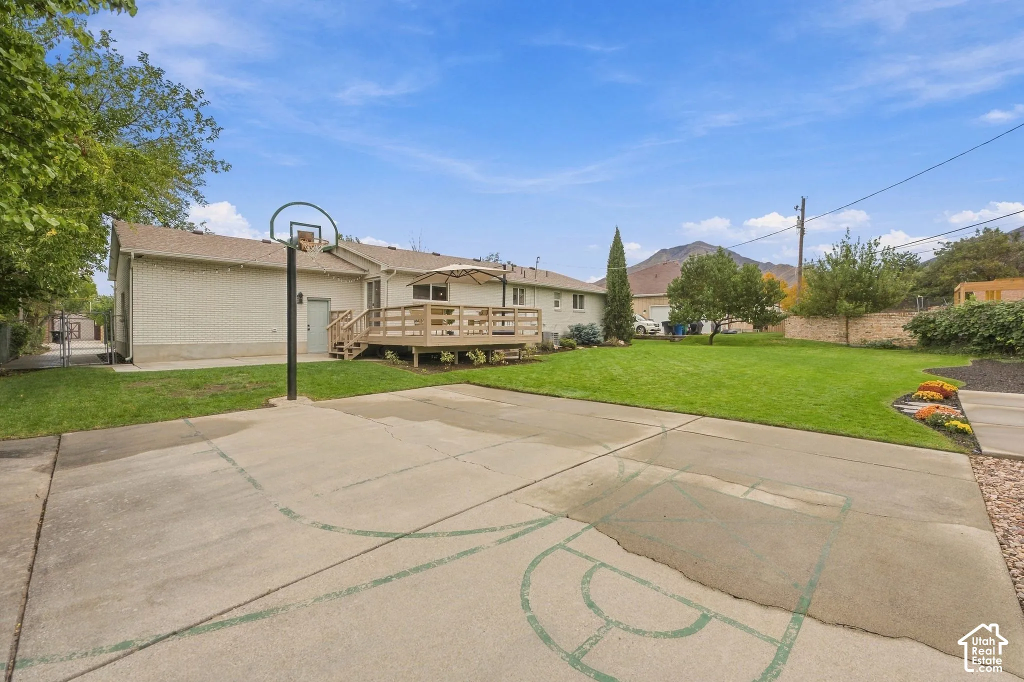 Exterior grounds featuring a concrete sports court with an integrated basketball hoop, a raised wood deck, expansive lawn, and mountain backdrop