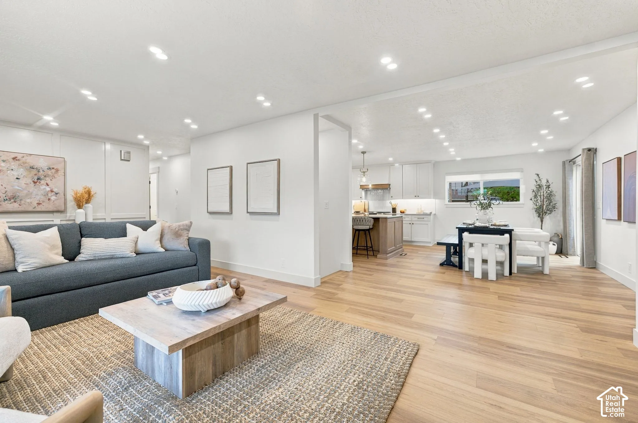 Open-concept interior featuring light wood-finish flooring, recessed lighting, white wall paneling, and kitchen cabinetry with a wood-toned island
