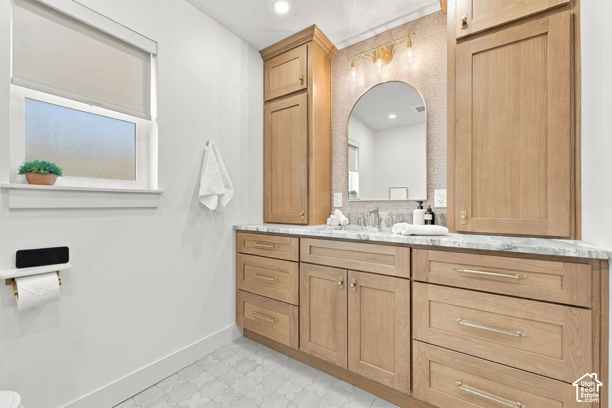 Vanity featuring natural finish wood cabinetry, stone-patterned countertop, arched wall mirror, integrated side towers, and patterned tile flooring