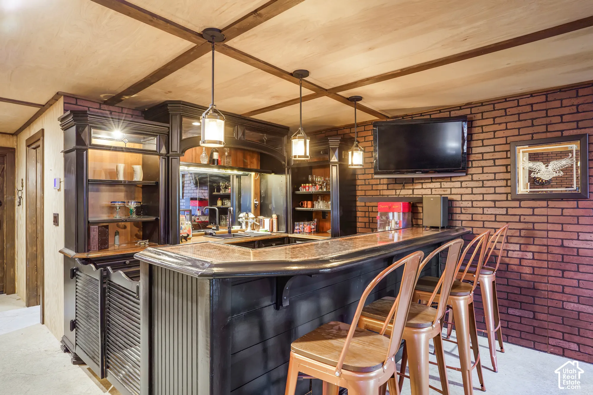 Custom built-in bar featuring dark cabinetry, metallic-finished countertop, exposed brick wall, wood-paneled ceiling with grid detailing, and trio of pendant light fixtures