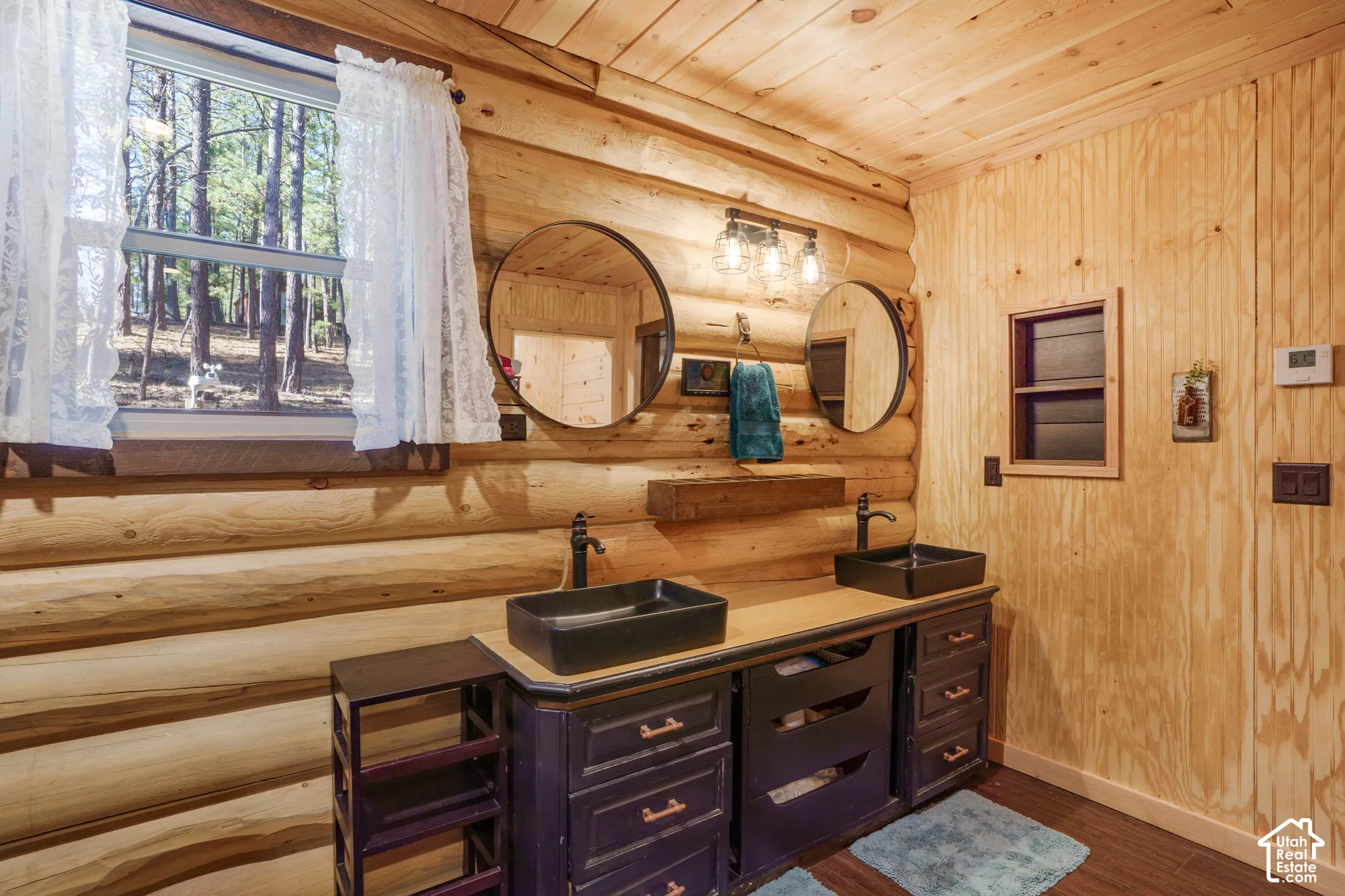 Bathroom featuring exposed log walls, dual black vessel sinks, dark-toned cabinetry, circular wall mirrors, and three-light vanity fixture