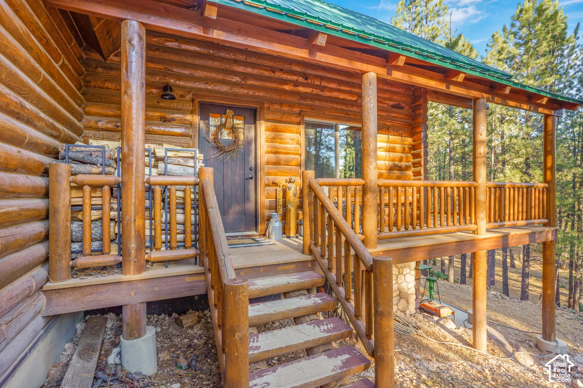 Log cabin exterior featuring a covered wraparound porch, natural log construction, green metal roof, and stone foundation masonry