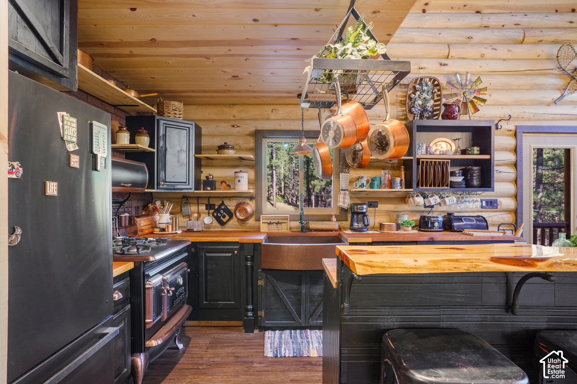 Log wall construction with exposed timber ceiling, copper farmhouse sink, custom dark-finished cabinetry, wood-slab island countertop, and professional-style range