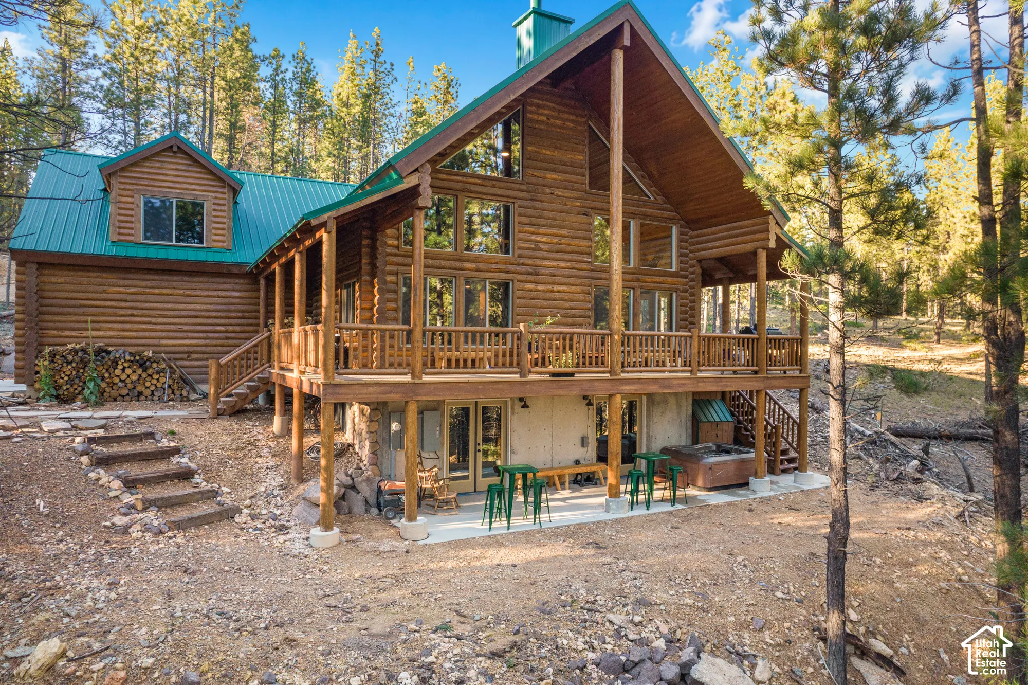 Log cabin exterior featuring a multi-level wood deck, green standing-seam metal roofing, large multi-pane windows, and a covered lower-level concrete patio