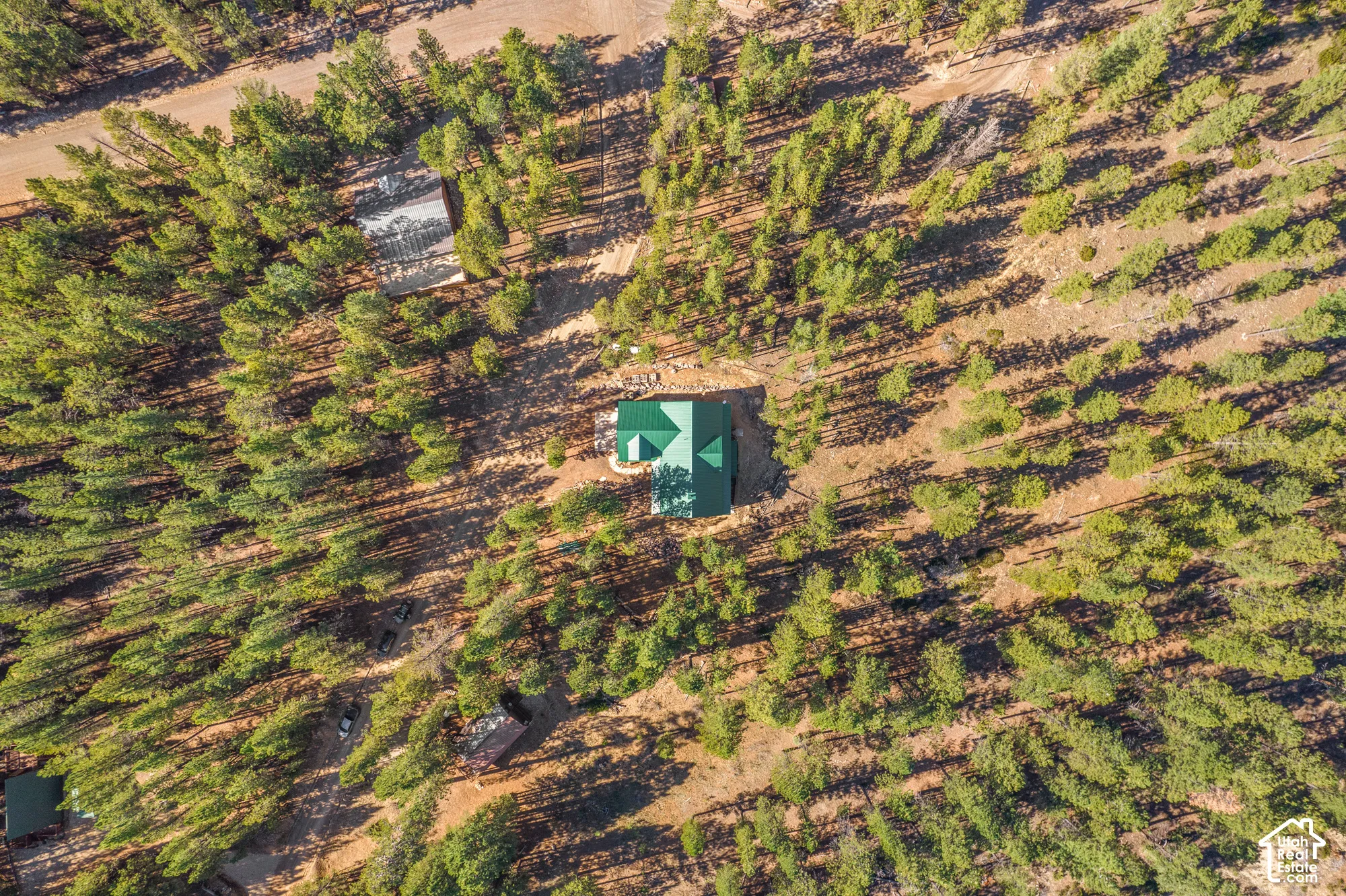 Aerial perspective of a detached structure featuring a multi-gable metal roof, situated on a large, heavily wooded lot with unpaved access paths