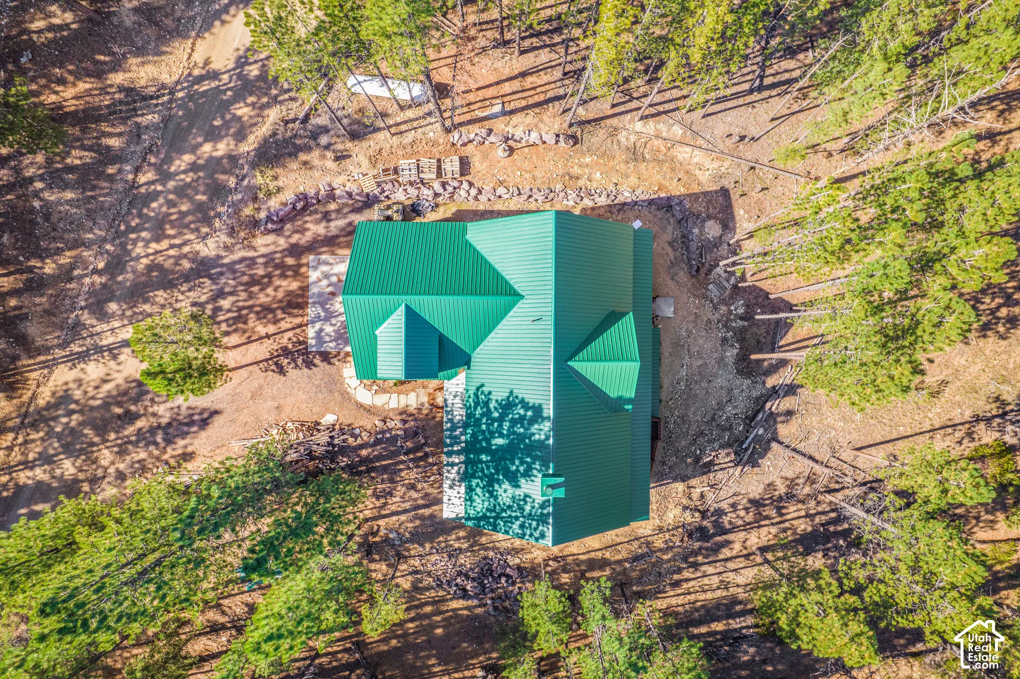 Aerial view of a structure featuring a green metal roof, stone-retaining walls, a concrete patio, and a wooded lot