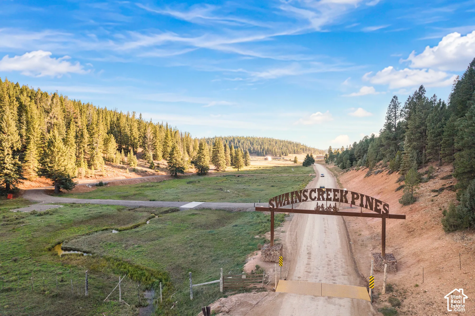 Swains Creek Pines entrance featuring a metal archway, stone-based pillars, dirt road, winding creek, and surrounding pine forest