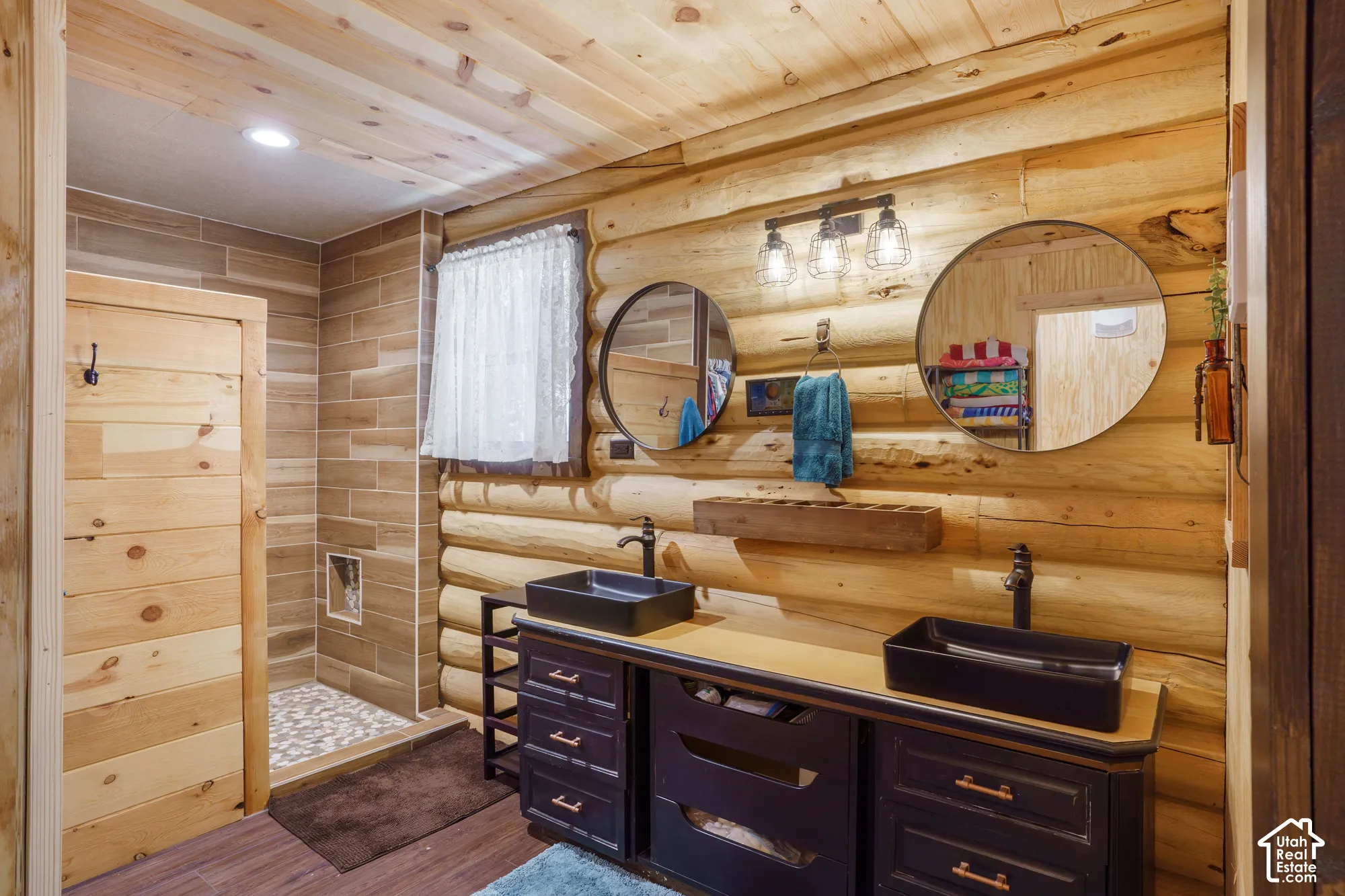 Log-style walls with matching wood-plank ceiling, double vanity with matte black rectangular vessel sinks, copper-finish hardware, and a walk-in shower featuring tiled walls and stone-texture flooring