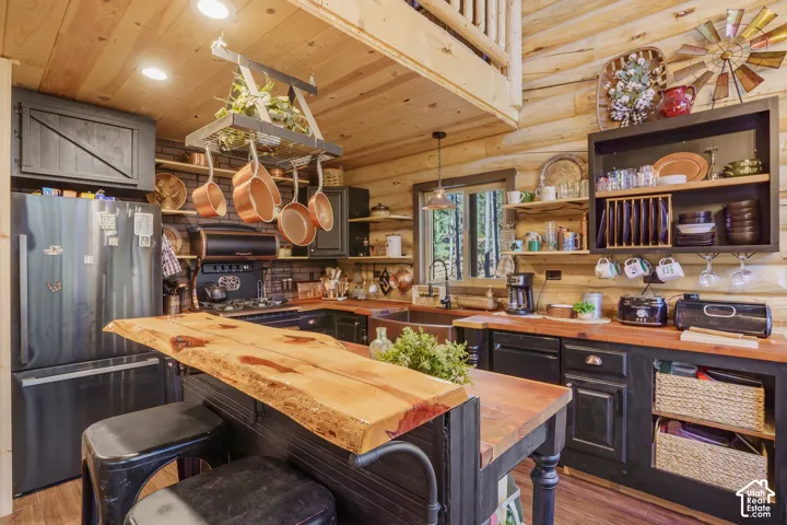 Rustic kitchen featuring exposed log walls, a live-edge wood island countertop, custom black cabinetry, a copper basin sink, and a suspended metal rack for cookware