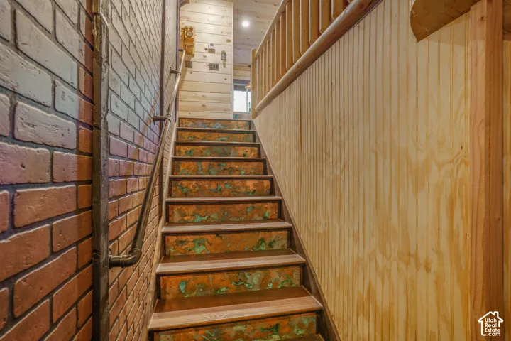 Staircase featuring custom metal risers, exposed brick wall, wood-finish treads, vertical wood paneling, and industrial-style pipe handrail