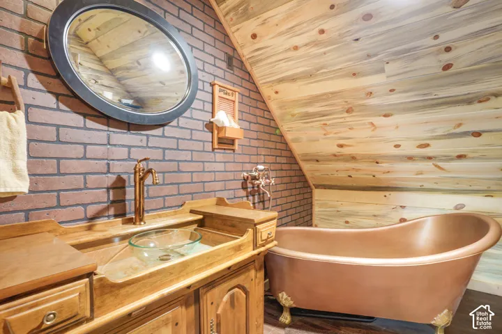 Brick-clad walls featuring a freestanding copper clawfoot tub, wood-finish sloped ceiling, vanity with a glass vessel sink, and metallic finish faucet