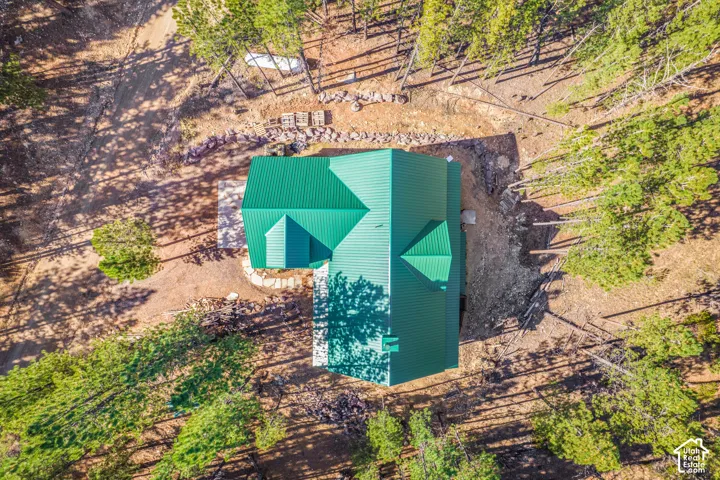 Aerial view of a structure featuring a green metal roof, stone-retaining walls, a concrete patio, and a wooded lot