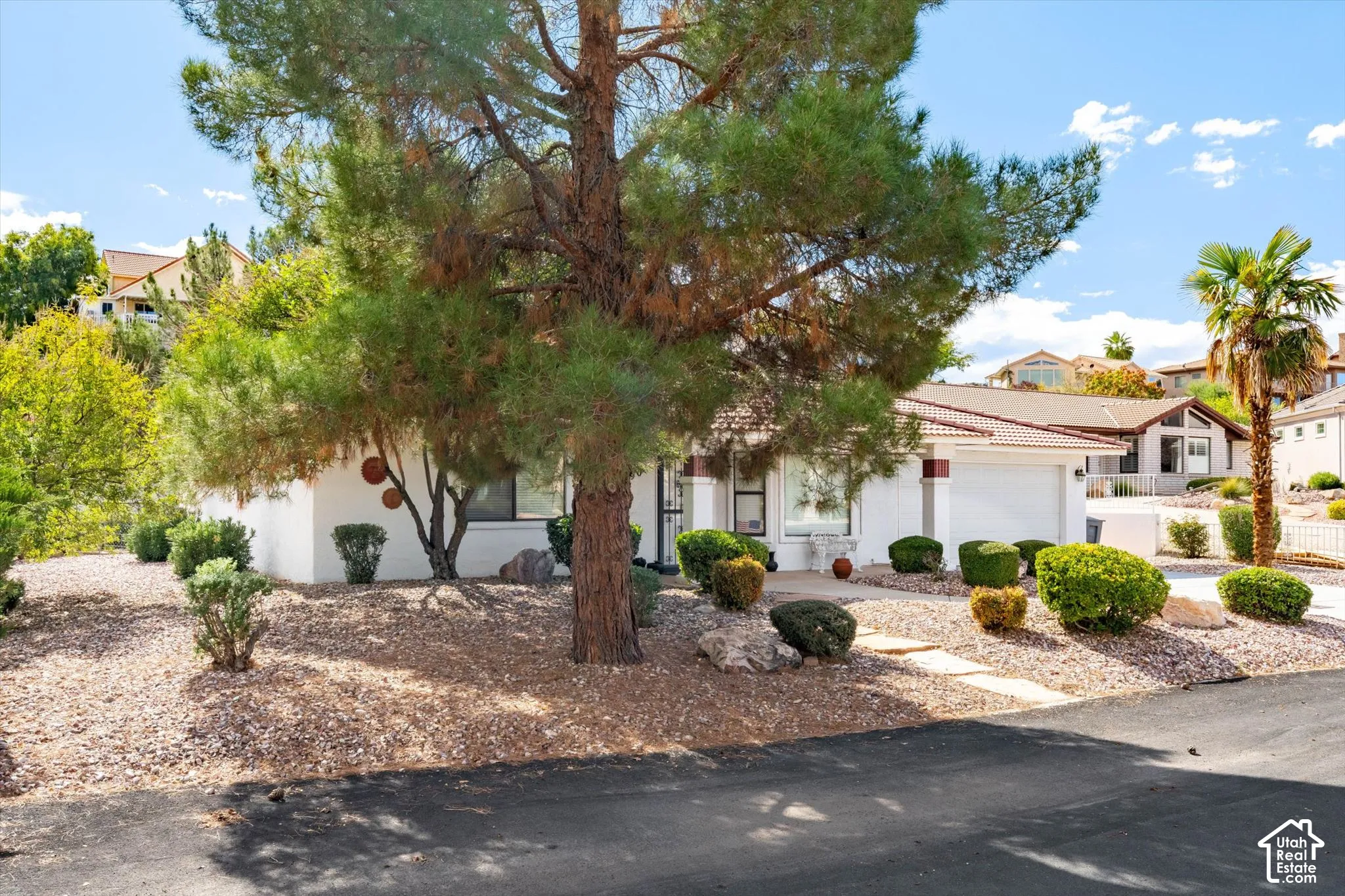 Exterior facade featuring a tile roof, light-toned stucco siding, attached two-car garage, mature pine tree, and xeriscaped front yard with stone path