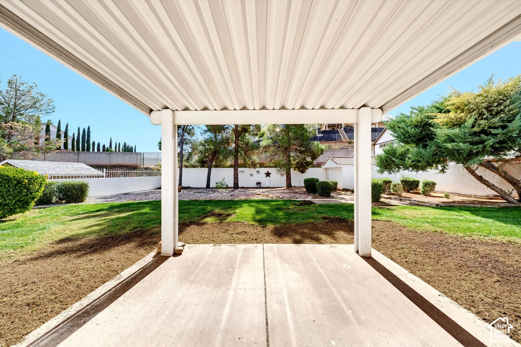 Covered concrete patio featuring a corrugated metal roof, manicured lawn, perimeter masonry wall, and mature landscaping