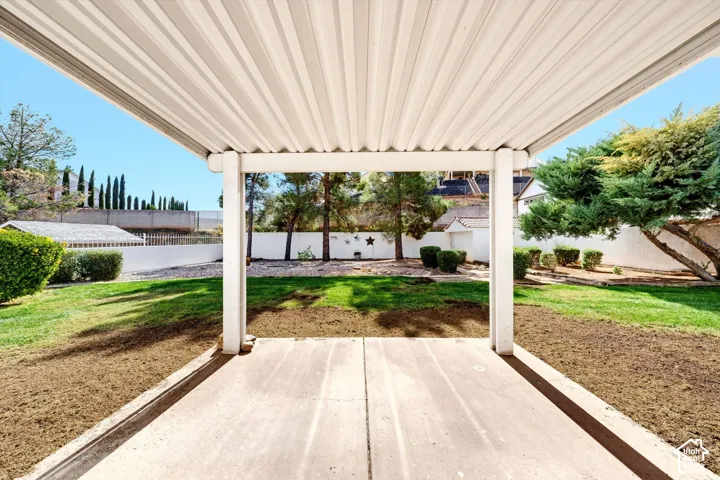 Covered concrete patio featuring a corrugated metal roof, manicured lawn, perimeter masonry wall, and mature landscaping
