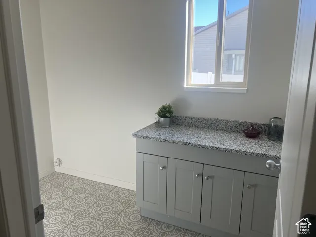 Utility area featuring patterned tile flooring, light gray shaker-style cabinetry, and stone-surface countertop, illuminated by a single-pane window