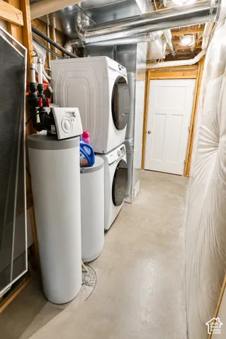 Utility area featuring a water softener system, stacked front-load laundry appliances, exposed metal ductwork, concrete flooring, and a white three-panel door