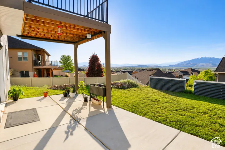 Covered concrete patio featuring an overhead deck with black metal railing, overlooking a lawn with perimeter fencing, corrugated metal raised garden beds, and mountain scenery