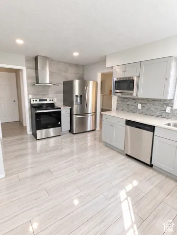 Kitchen featuring light gray cabinetry, stainless steel appliances, a modern chimney-style range hood, decorative tile backsplash, and wood-finish flooring