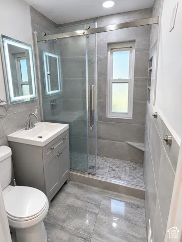 Bathroom featuring large-format gray tile flooring and wall surfacing, walk-in shower with pebble-stone flooring, frameless glass sliding door, and a wall-mounted LED-backlit mirror