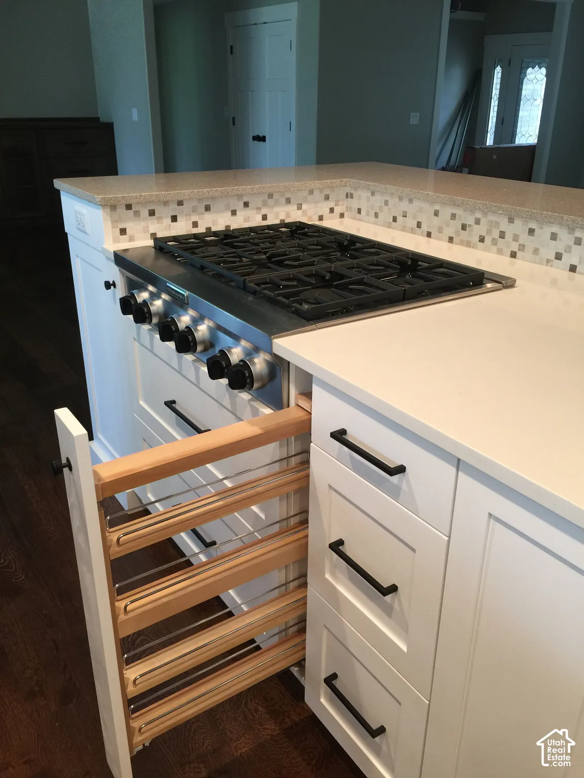 Kitchen island featuring a gas cooktop, stone-finish countertops, glass-tile backsplash, white shaker-style cabinetry with matte black hardware, and a built-in pull-out spice rack