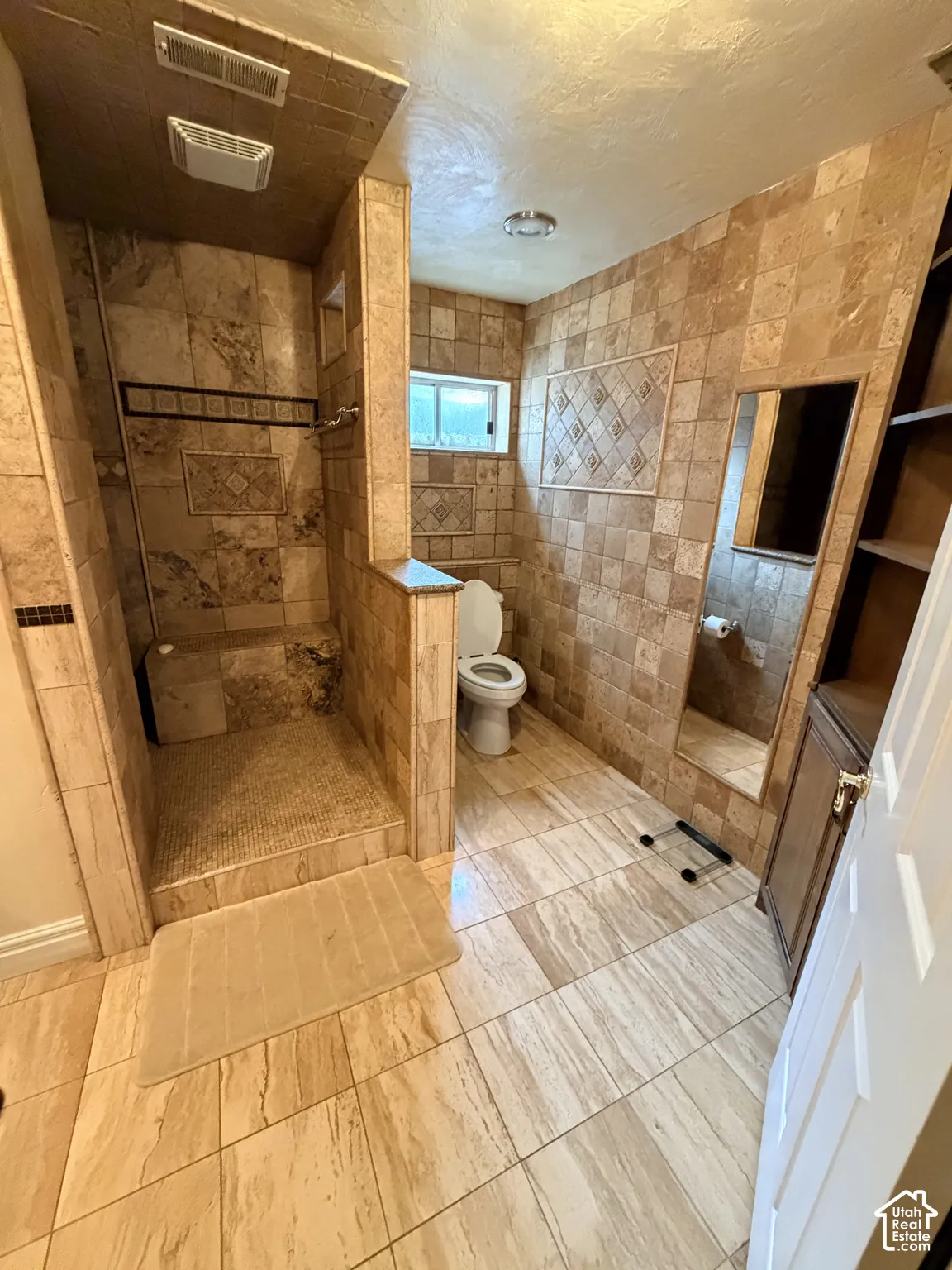 Full-wall stone tile surround, walk-in shower with integrated bench seat, inset decorative tile accents, built-in shelving, and floor-to-ceiling mirror