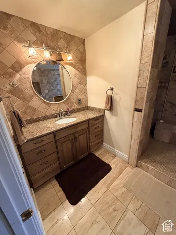 Bathroom featuring a stained-wood vanity with stone-finish countertop, diagonal wall tile backsplash, circular wall-mounted mirror, three-light fixture, and large-format floor tiling