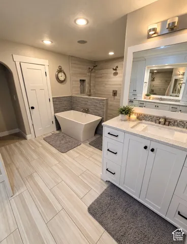 Bathroom featuring wood-finish tile flooring, a freestanding soaking tub, a tiled walk-in shower with a built-in niche, white shaker-style cabinetry with quartz countertops, and a modern vanity mirror with integrated lighting