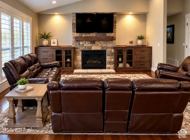 Floor-to-ceiling stone fireplace with a thick timber mantel, built-in custom wood cabinetry with glass-paneled doors, wood-finish flooring, vaulted ceiling, and plantation shutters