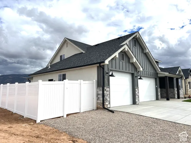 Multi-textured exterior featuring stucco, vertical siding, and stone accents, two-car garage with overhead lighting, and white vinyl privacy fencing