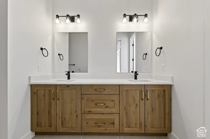 Double vanity featuring wood-finish cabinetry with brass-tone hardware, white stone-composite countertop, dual undermount sinks with matte black faucets, and dual wall-mounted light fixtures