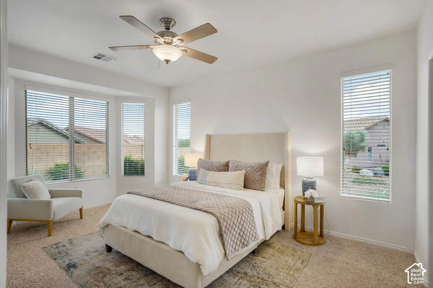 Neutral wall-to-wall carpeting, multiple large windows, recessed bay window area, and a ceiling-mounted light fixture with fan