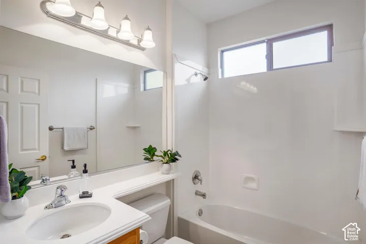 Bathroom featuring a white vanity with a single basin sink, wall-spanning mirror, four-light fixture, and a white tub-shower combination with a horizontal window