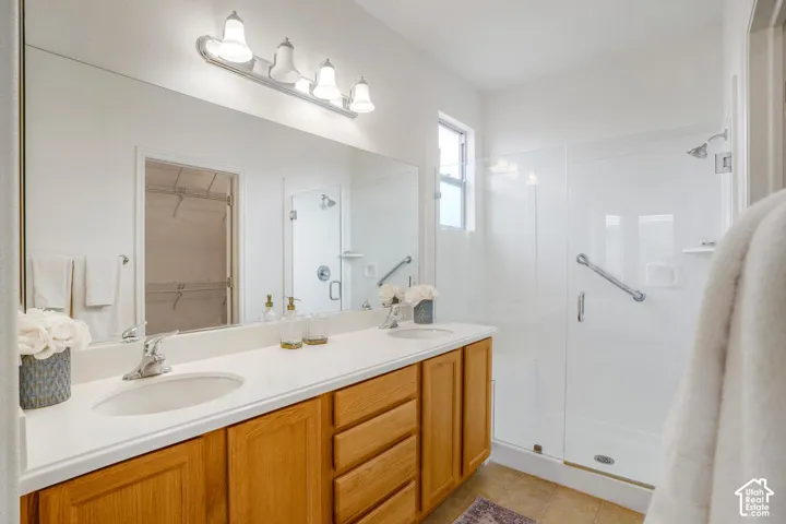 Double-sink vanity with natural wood-finish cabinetry, solid-surface countertop, full-width vanity mirror, four-light fixture, and walk-in shower with glass enclosure