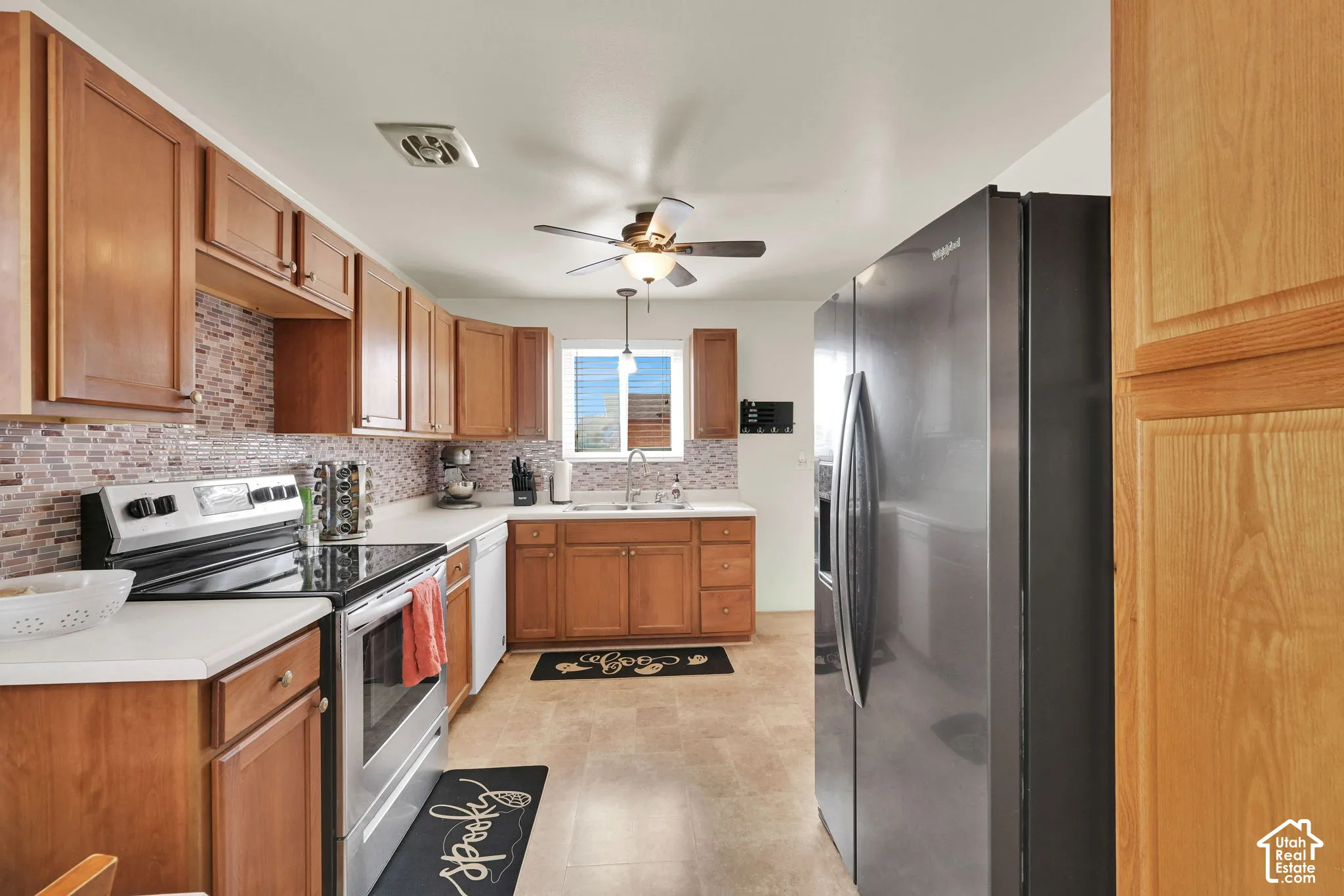 Kitchen featuring natural-finish wood cabinetry, mosaic tile backsplash, solid-surface countertops, integrated ceiling fan with light fixture, and stainless steel appliances