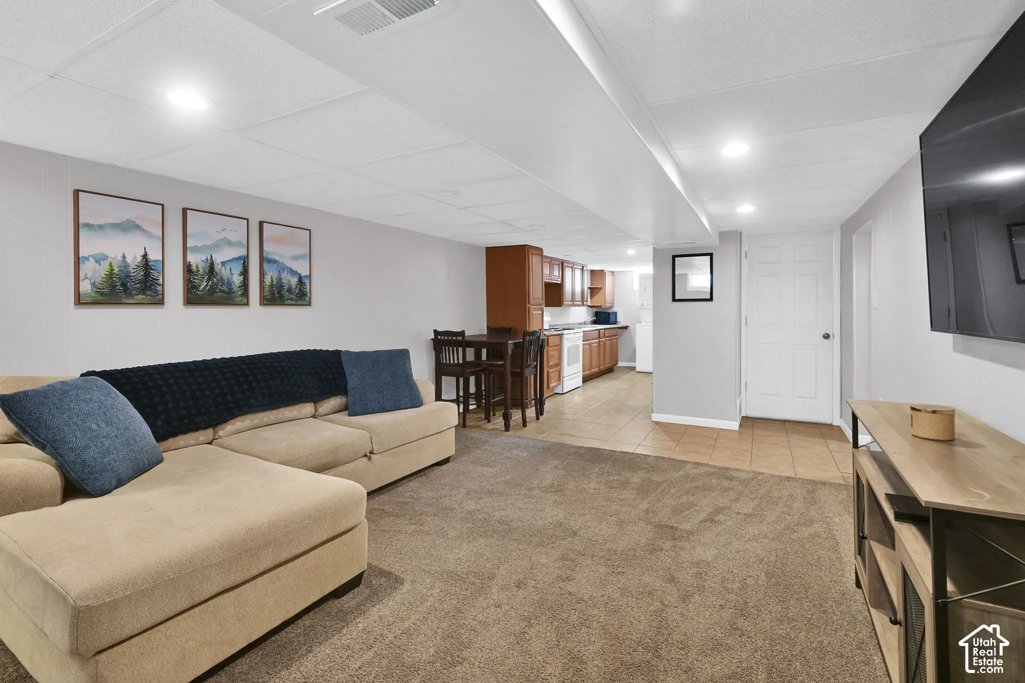 Finished room featuring recessed lighting, a drop ceiling, wall-to-wall carpeting, and a kitchen area with wood cabinetry and tile flooring