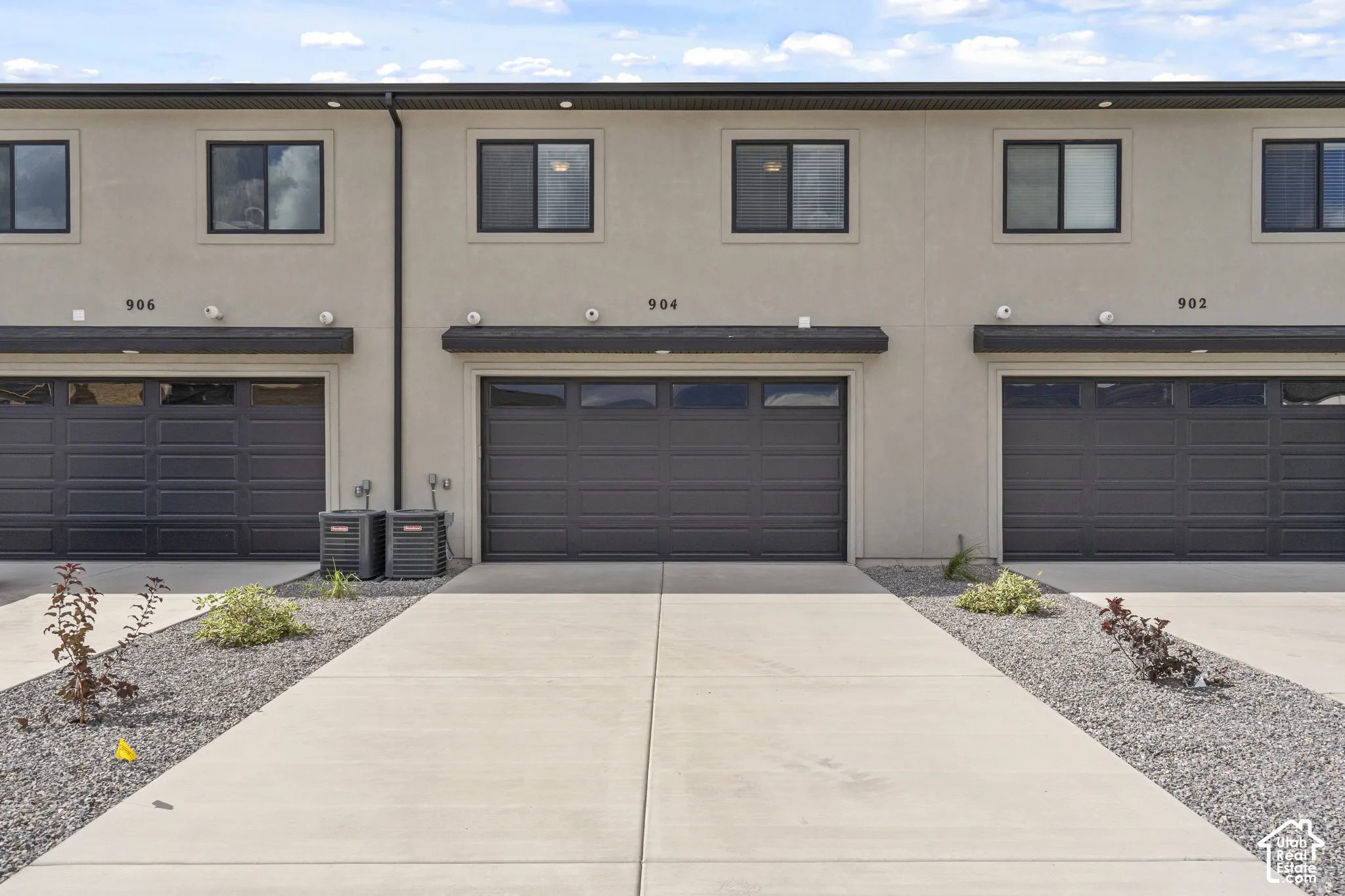 Multi-unit exterior featuring a smooth stucco facade, dark-toned garage doors with inset windows, concrete driveways, and stone-ground landscaping