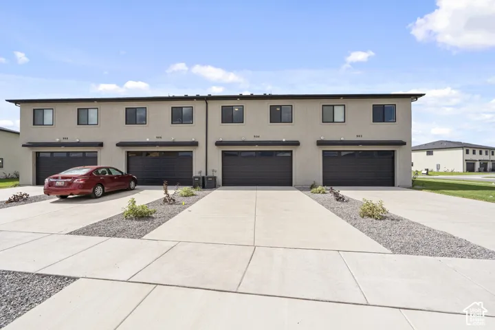Two-story townhome exterior featuring smooth stucco siding, four individual attached garages with dark-finished doors, concrete driveways, and xeriscaped front yard accents