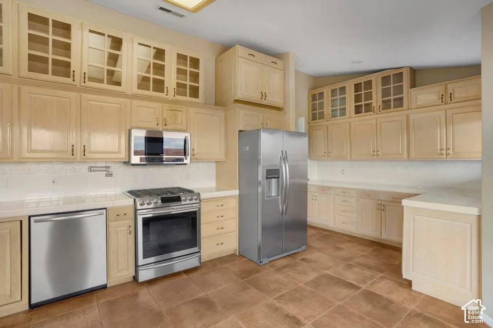 Kitchen featuring light wood cabinetry with glass-panel upper doors, stainless steel appliances, tile countertops, and ceramic tile flooring