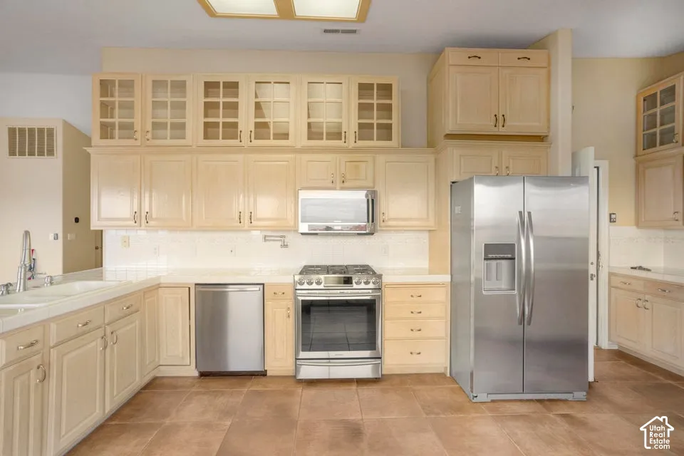 Kitchen featuring light-tone cabinetry with glass-paneled upper doors, tile flooring, stainless steel appliances, and a tiled backsplash