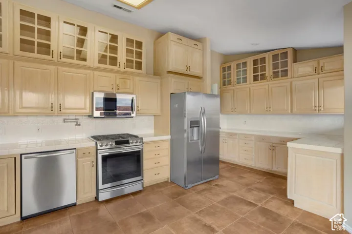 Kitchen featuring light wood cabinetry with glass-panel upper doors, stainless steel appliances, tile countertops, and ceramic tile flooring