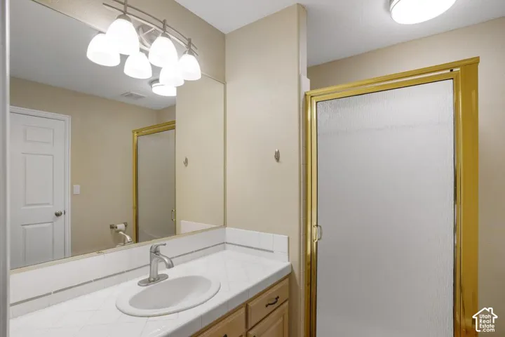 Bathroom vanity featuring a tiled countertop with an integrated basin, wooden cabinetry, a large wall-mounted mirror, multi-light fixture, and a shower enclosure with a metal frame