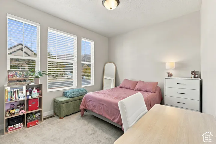 Neutral-toned bedroom featuring three large windows with horizontal blinds, textured carpeting, and a flush-mount ceiling light fixture