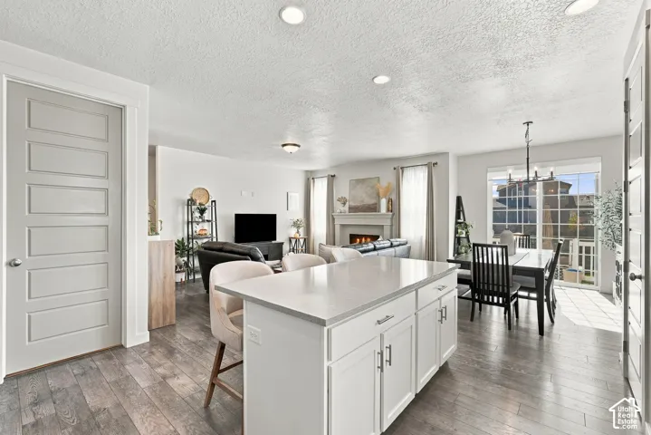 Kitchen island featuring a solid-surface countertop and shaker-style cabinetry, complemented by wood-finish flooring, recessed lighting, and a fireplace with a decorative mantel