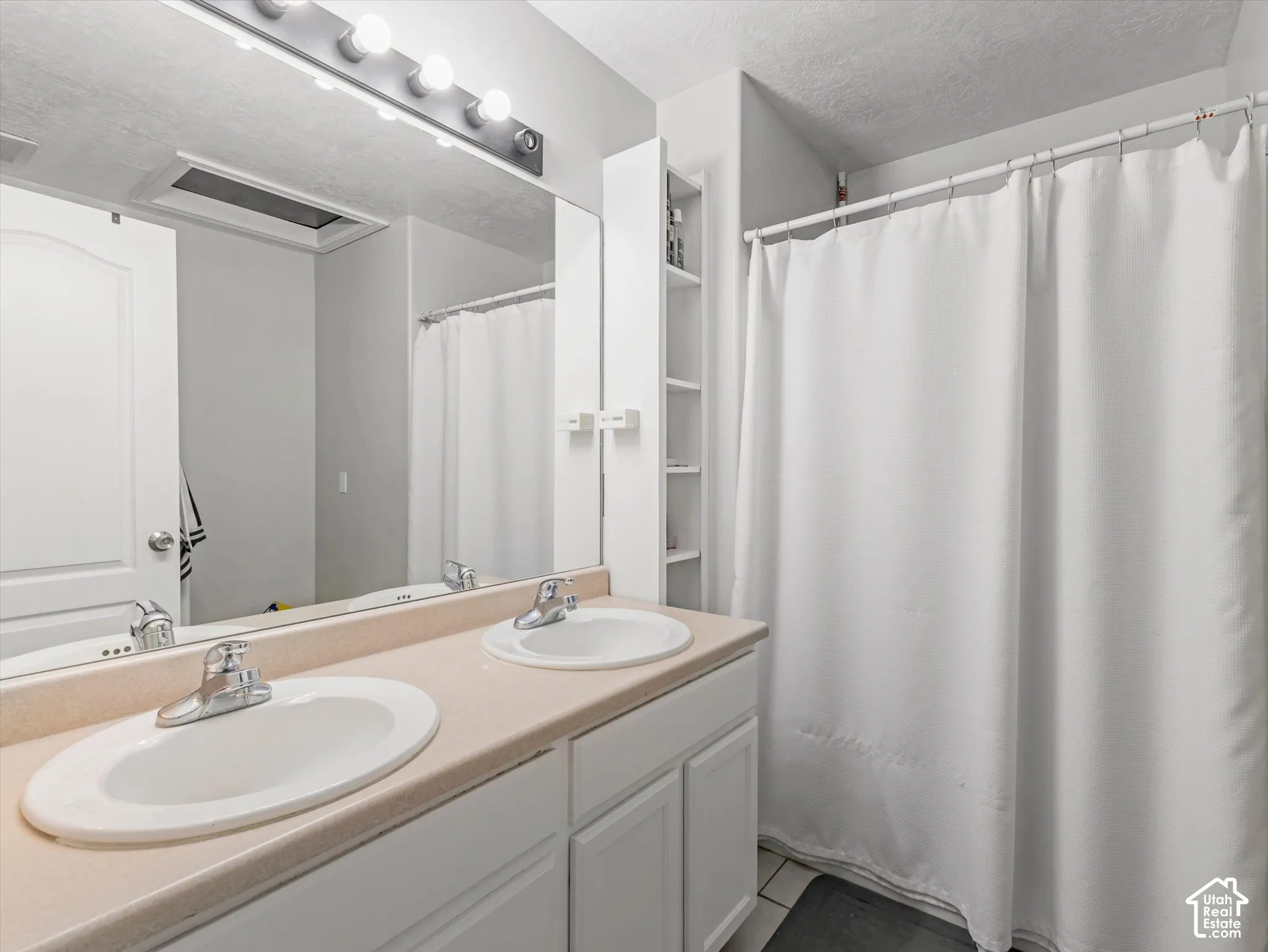 Dual-sink vanity with a solid-surface countertop, white cabinetry, integrated shelving, and a large-format vanity mirror with an overhead light fixture
