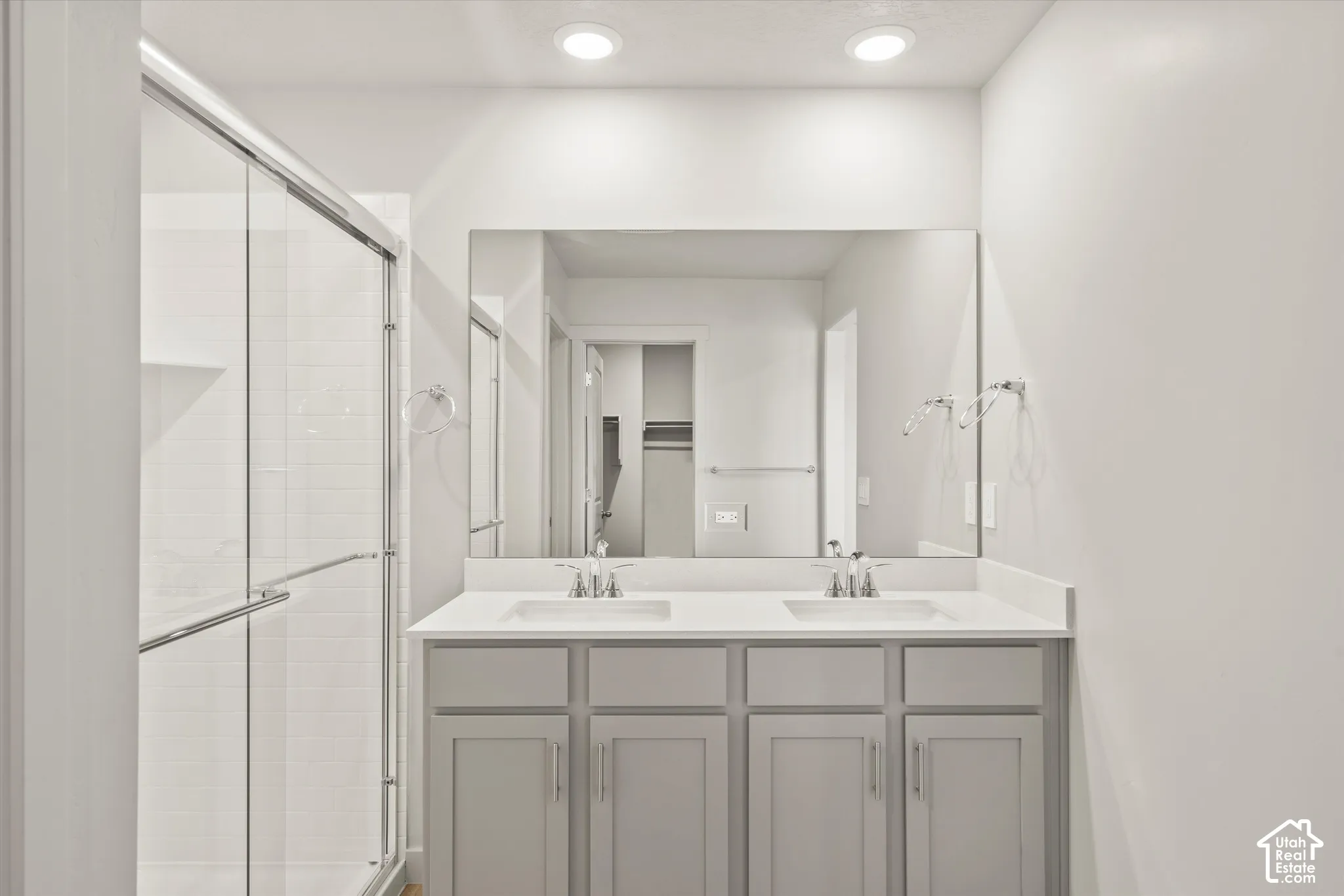 Dual-sink vanity with solid-surface countertop, cabinetry featuring brushed nickel hardware, oversized wall-mounted mirror, and glass-enclosed shower with tile surround