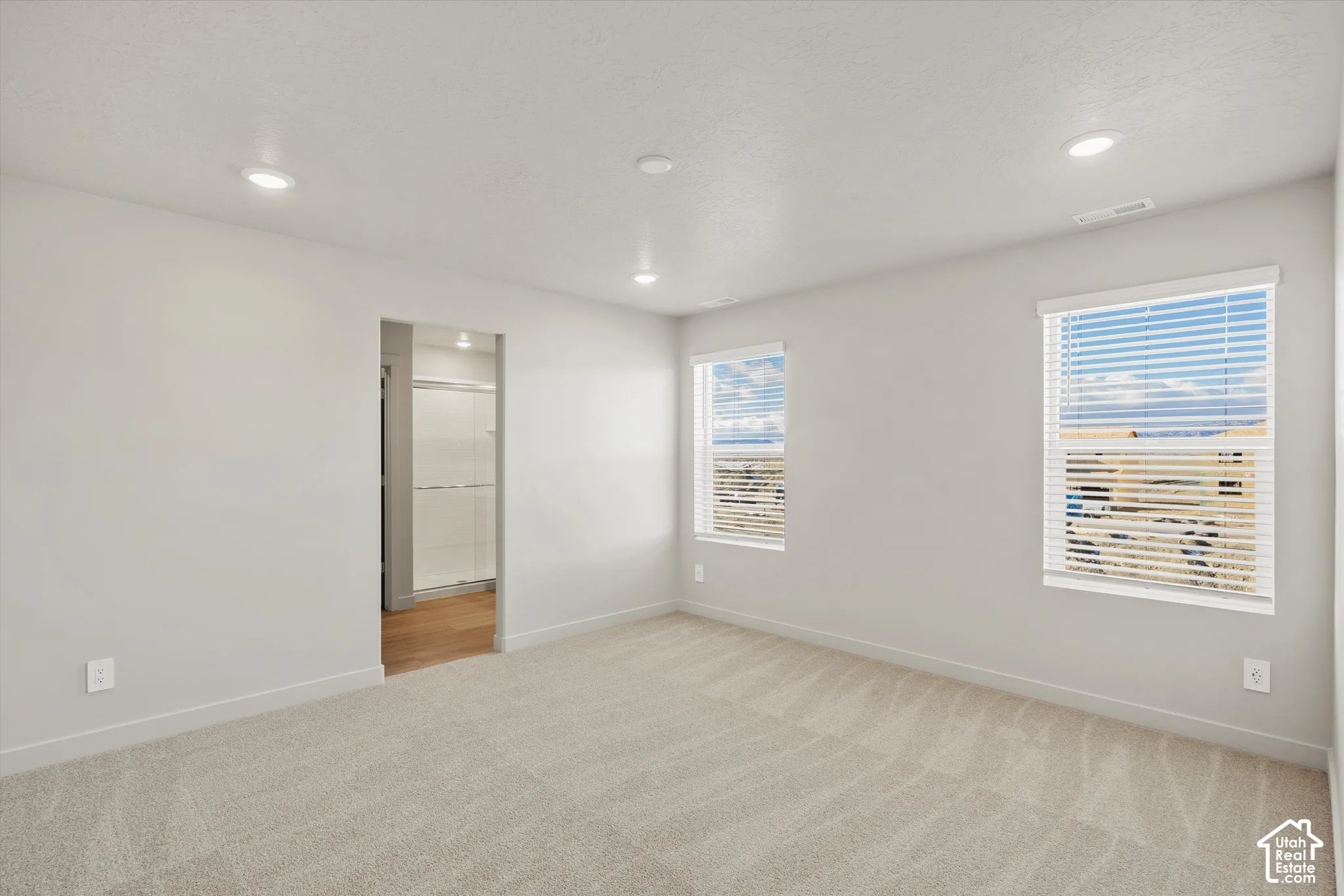 Room featuring neutral carpeting, recessed lighting, dual windows with horizontal blinds, and a doorway revealing secondary space with hard-surface flooring