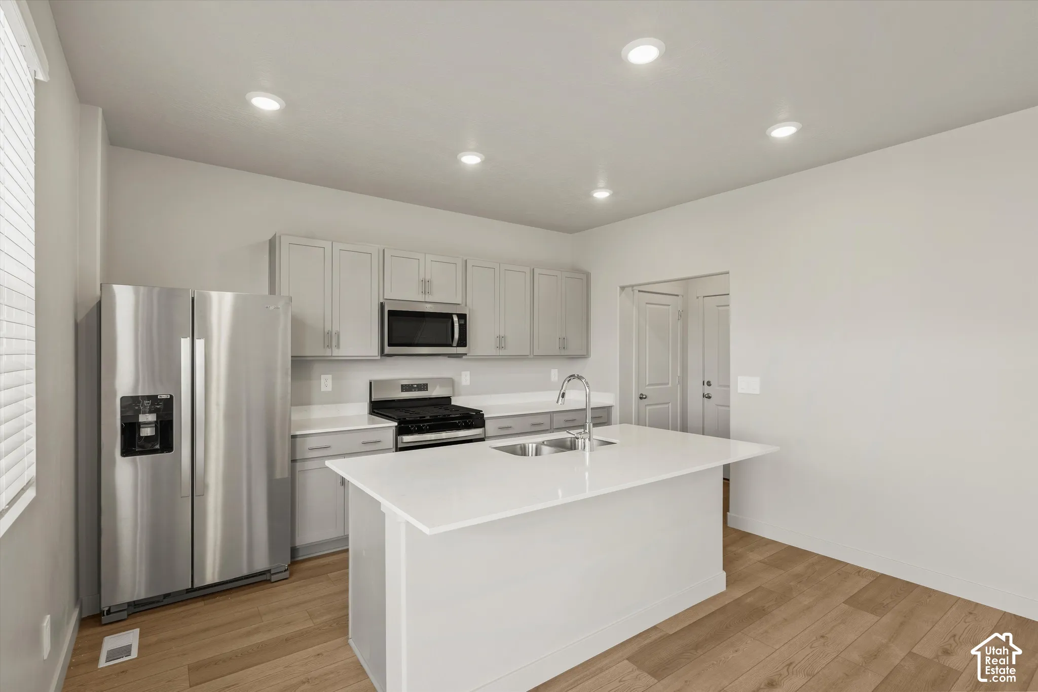 Kitchen featuring light grey cabinetry, quartz countertops, a central island with a dual-basin sink, stainless steel appliances, and wood-finish flooring