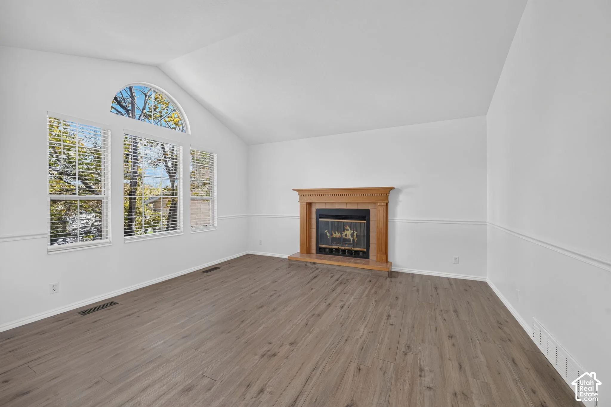 Vaulted ceiling living area featuring wood-finish flooring, a wood-surround fireplace with a brick hearth, and an arched window configuration